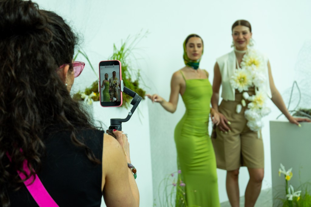 A woman taking a photo of two models posing in elegant outfits among decorative plants.