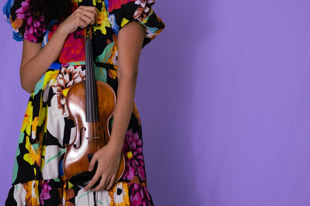A musician in a colorful floral dress holds a violin while standing against a purple background.