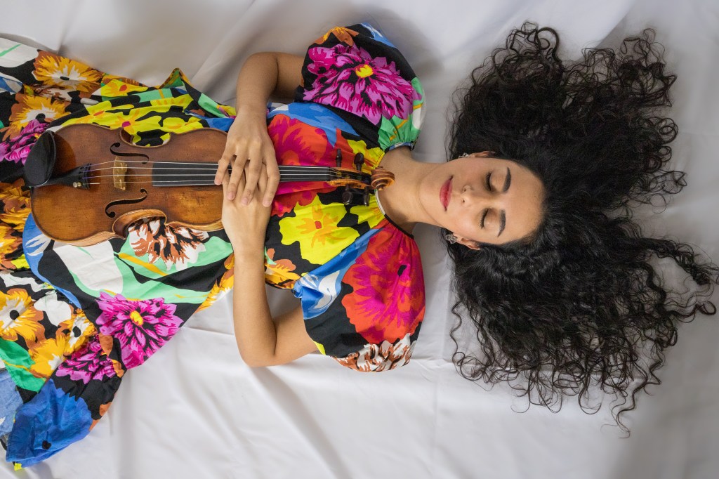 A musician and violinist, lying on a white surface, wearing a colorful floral dress and holding her violin, with her curly hair spread out.