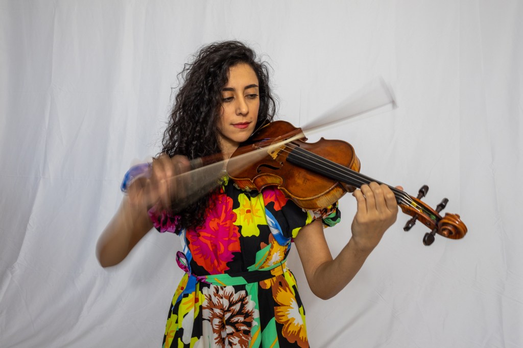 Ava Shadmani playing the violin, wearing a colorful floral dress against a plain white background.