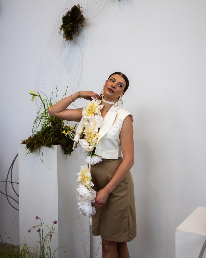 A model wearing a white sleeveless top and beige skirt poses with a floral necklace in front of a minimalist background featuring greenery.