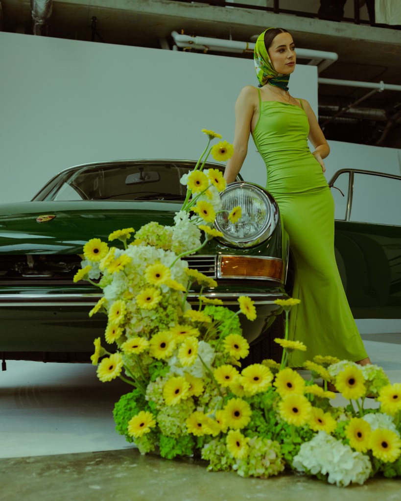 A woman in a green dress and headscarf poses next to a classic green car, surrounded by colorful flowers.